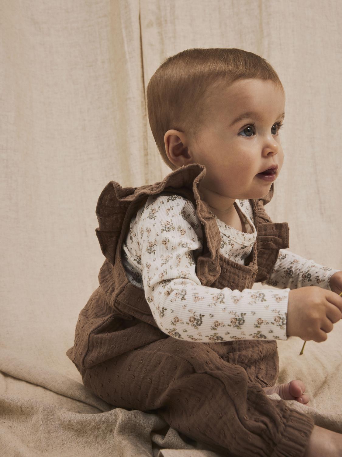 Söt liten flicka klädd i bruna hängselbyxor med volanger och en body till baby flicka, vit body med små gulliga blommor. Bebis body i ekologisk bomull.