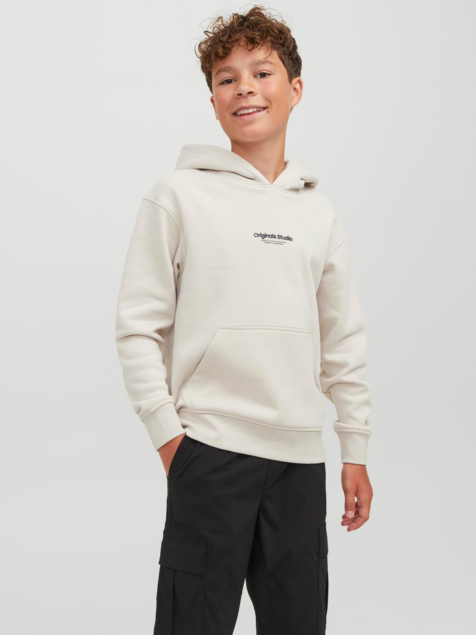 Kille klädd i Vit, naturvit hoodie från Jack & Jones till ungdom kille, pojke. Luvtröja i enfärgat vitt med logga på bröstet i deras omtyckta modell Vesterbro. FIcka på magen.