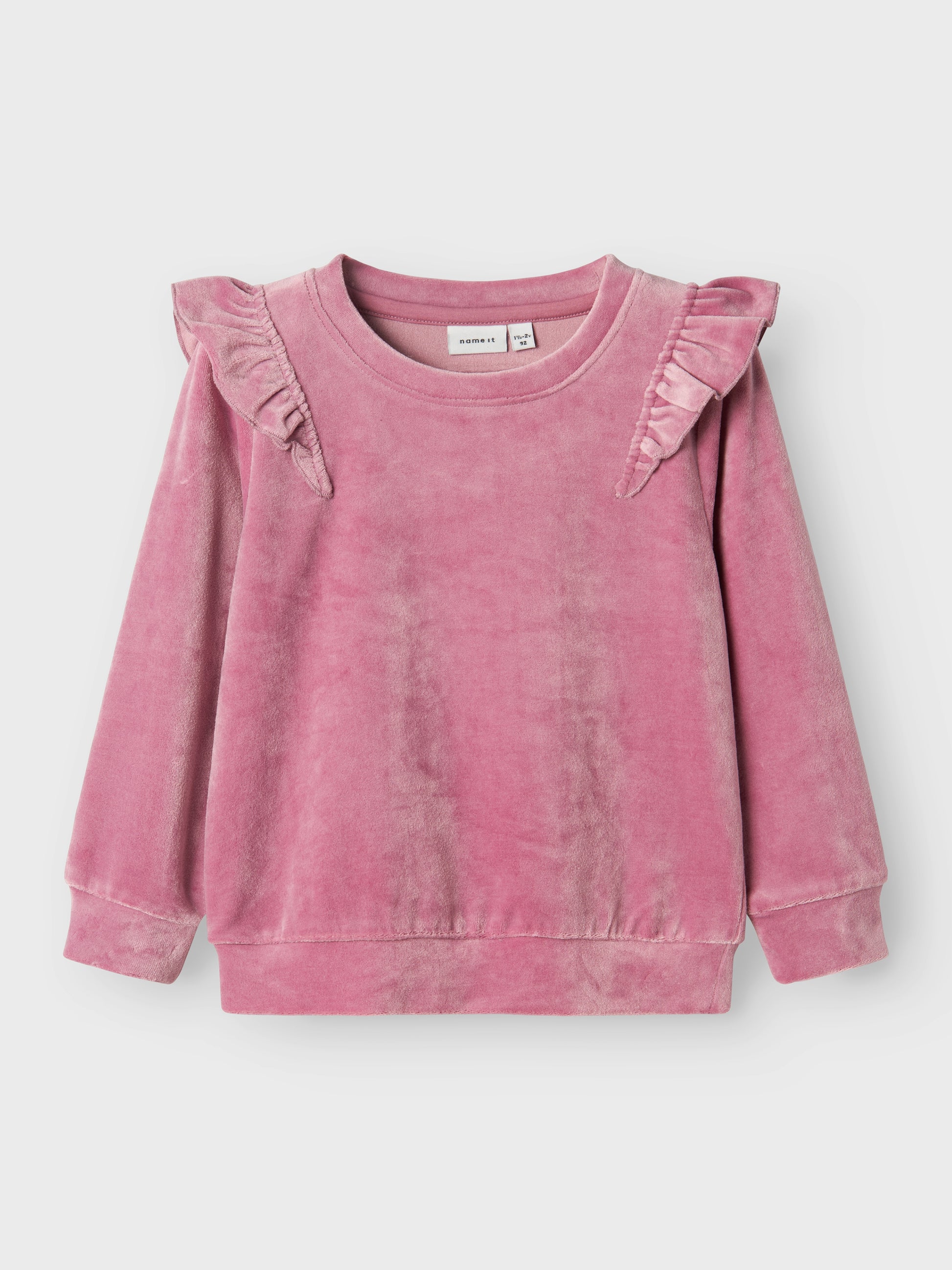 Rosa mysig velour tröja till barn med volanger på axlarna. Låt din flicka få en söt mjuk tröja i återvunnen polyester. 
Välkommen bekväma och coola sweatshirts till ditt barns garderob och skapa en trendig och avslappnad look. Velour är ett mjukt och glänsande tyg med fibrer som följer handens drag för en fin effekt.

Produkttyp: Sweatshirts
Hals: Rund hals
Ärm: Lång ärm
Muddar: Ribbstickade muddar
Passform: Normal passform